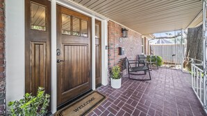 A brick house with a wooden front door, a covered porch with two chairs, and a welcome mat. - 3BR/3BA 1940s Bungalow Near Expo Square. Pet friendly! (Tulsa)