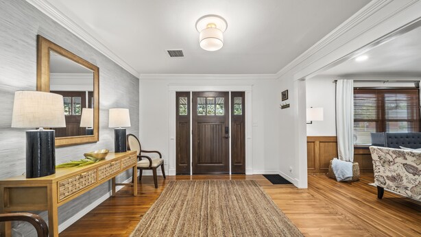 A spacious entryway with a wooden floor, a dark wooden door, a mirror, a console table with lamps, and a patterned rug. - 3BR/3BA 1940s Bungalow Near Expo Square. Pet friendly! (Tulsa)