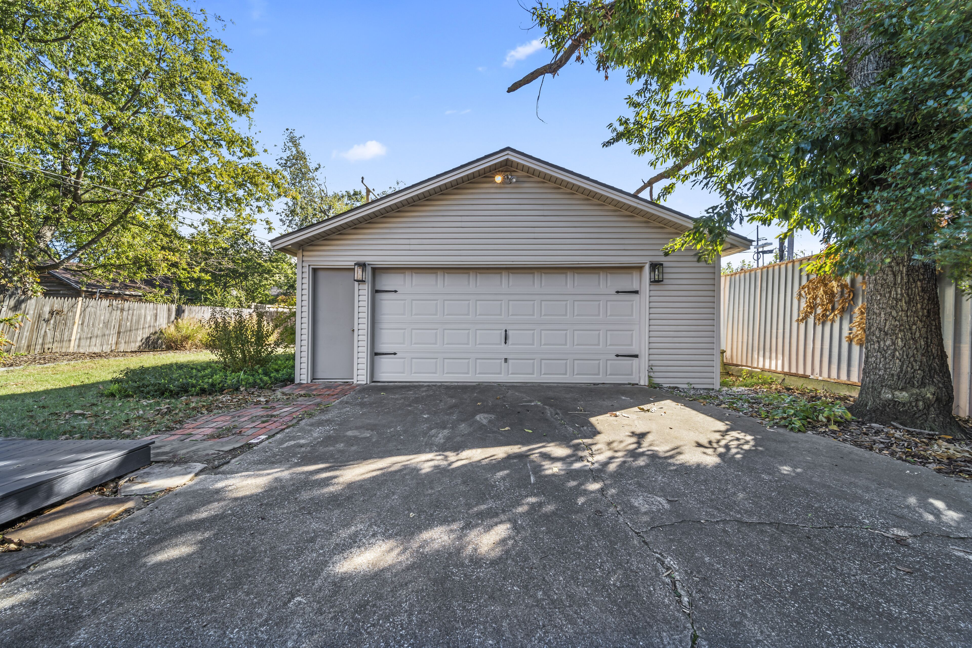 Un garage de plain-pied avec une porte blanche, entouré d’une clôture en bois, d’arbres et d’une allée pavée.