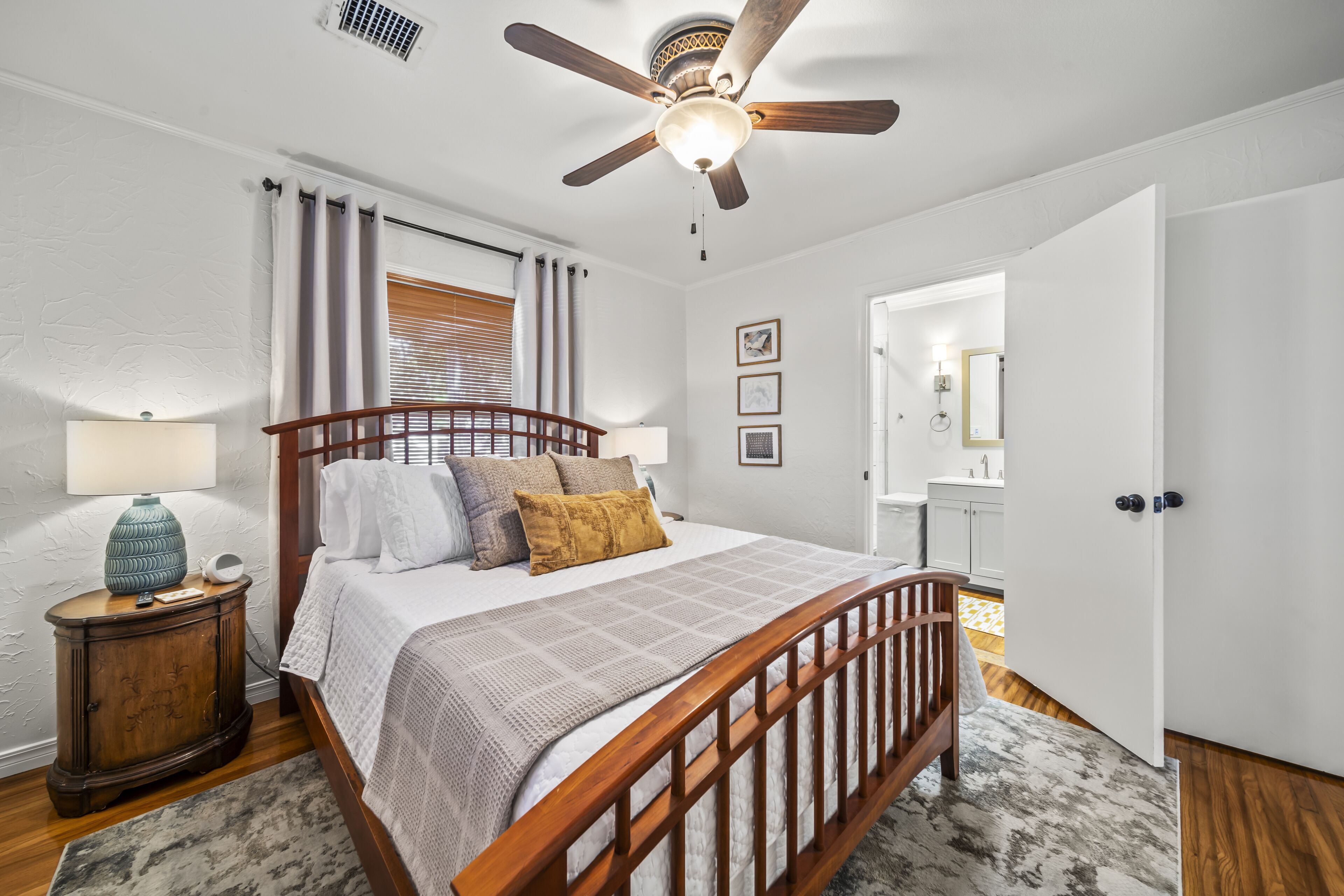 A bedroom with a wooden bed, a ceiling fan, a nightstand with a lamp, and a bathroom visible through an open door.