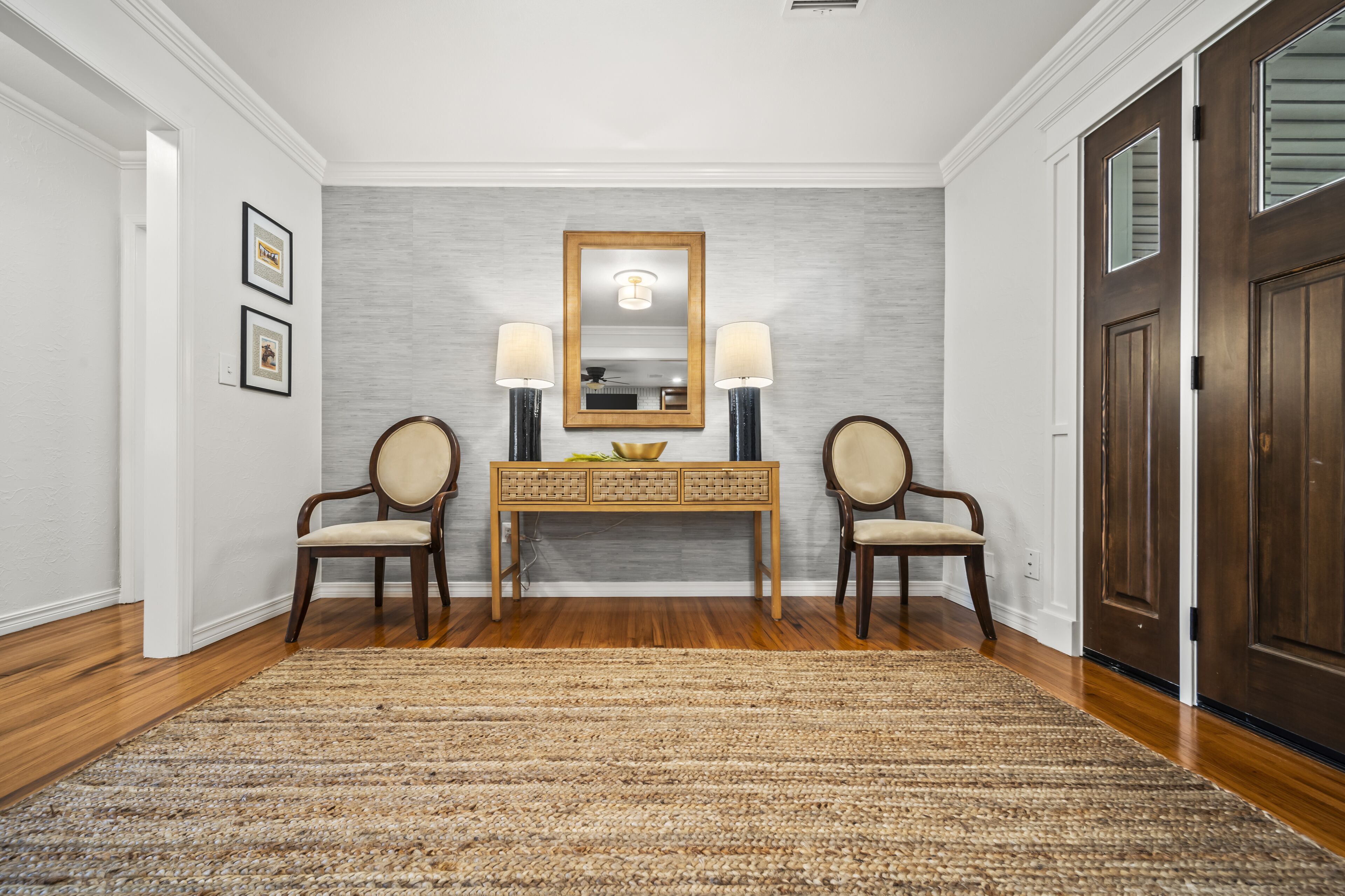 A hallway with a wooden floor, a beige rug, a wooden console table with a mirror, two chairs, and a dark wooden door.