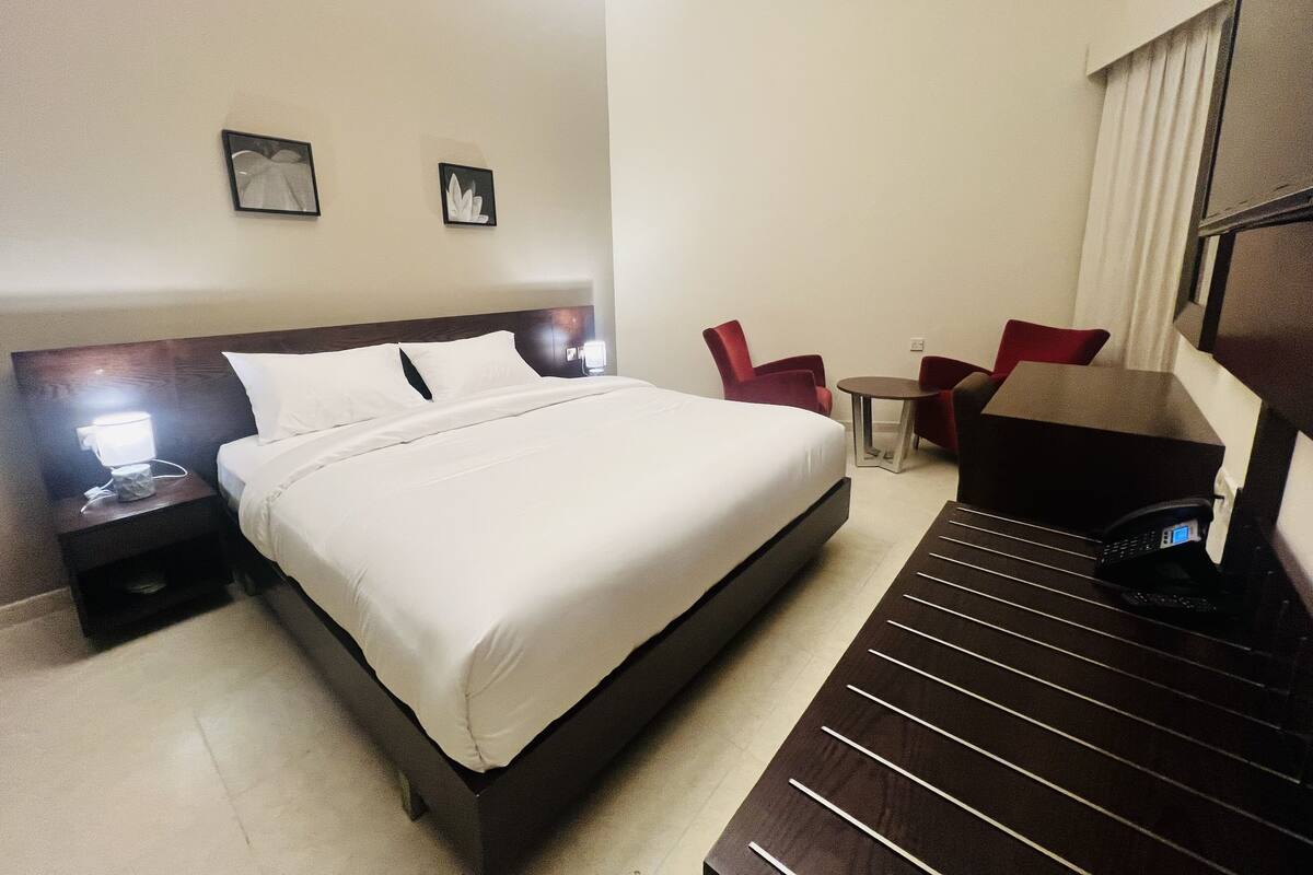 Ein Hotelzimmer mit einem Bett, einem Schreibtisch, zwei Stühlen, einem kleinen Tisch und zwei an der Wand befestigten Bildern.