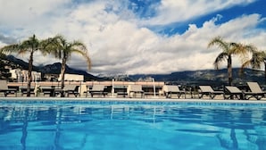 Piscina con sillones de playa y palmeras, con montañas y un cielo parcialmente nublado al fondo.