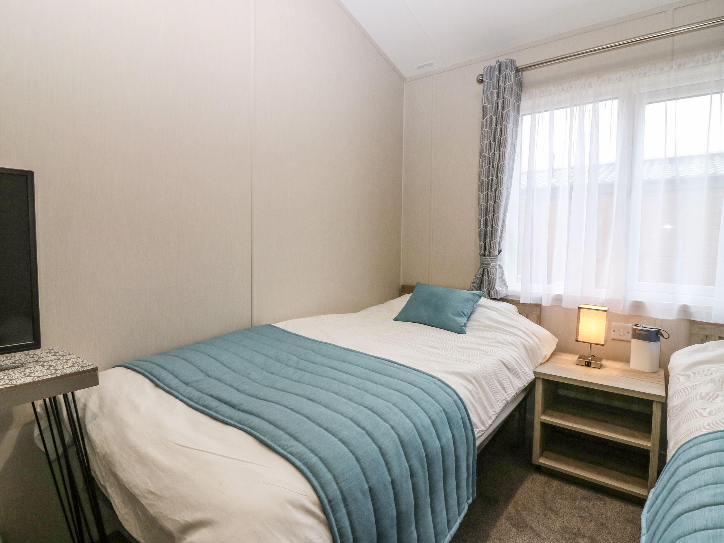 A bedroom with a bed, a bedside table with a lamp, a TV on the wall, and a window with curtains.