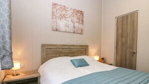 A neatly made bed with a wooden headboard, a blue pillow, and a blue blanket. There is a bedside table with a lamp and a small speaker. A painting hangs on the wall above the bed.
