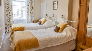 A bedroom with two beds, a window with curtains, a bedside table with a lamp, and a wooden wardrobe. - Victoria House (Alnwick)