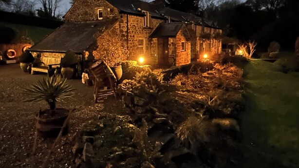 A stone building with a sloped roof, surrounded by a garden with various plants and a small pond. - Fiddlers Green Cottage, Tenby (Saint Florence)