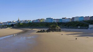 A sandy beach with a large rock formation and a person walking. In the background, there are colorful buildings along the coastline. - Fiddlers Green Cottage, Tenby (Saint Florence)