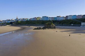 A sandy beach with a large rock formation and a person walking. In the background, there are colorful buildings along the coastline.