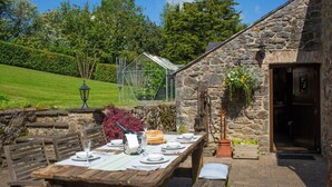 A stone building with a wooden table set for dining, a glass greenhouse, and a stone wall. - Fiddlers Green Cottage, Tenby (Saint Florence)