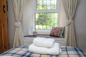A bed with a plaid bedspread, a window with curtains, a small bedside table with a clock, and a colorful pillow.