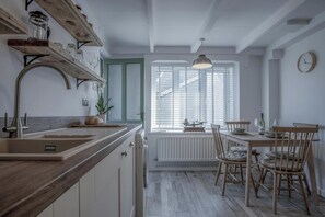 A kitchen with a wooden countertop, a sink, a dining table with chairs, a window with blinds, and a hanging light fixture.