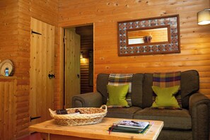 A cozy cabin interior with a wooden wall, a woven basket on a table, and a framed mirror above a couch with green cushions. - Fiddlers Green Log Cabin, Tenby (Saint Florence)
