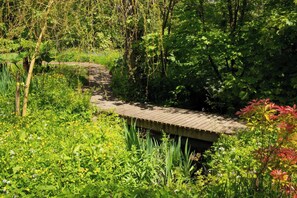 A wooden bridge over a stream in a lush garden with dense foliage and various plants.
