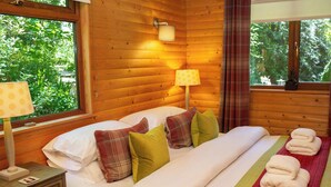 A wooden cabin bedroom with a bed, pillows, a nightstand, a lamp, and a window with curtains. - Fiddlers Green Log Cabin, Tenby (Saint Florence)