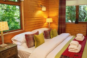 A wooden cabin bedroom with a bed, pillows, a nightstand, a lamp, and a window with curtains.