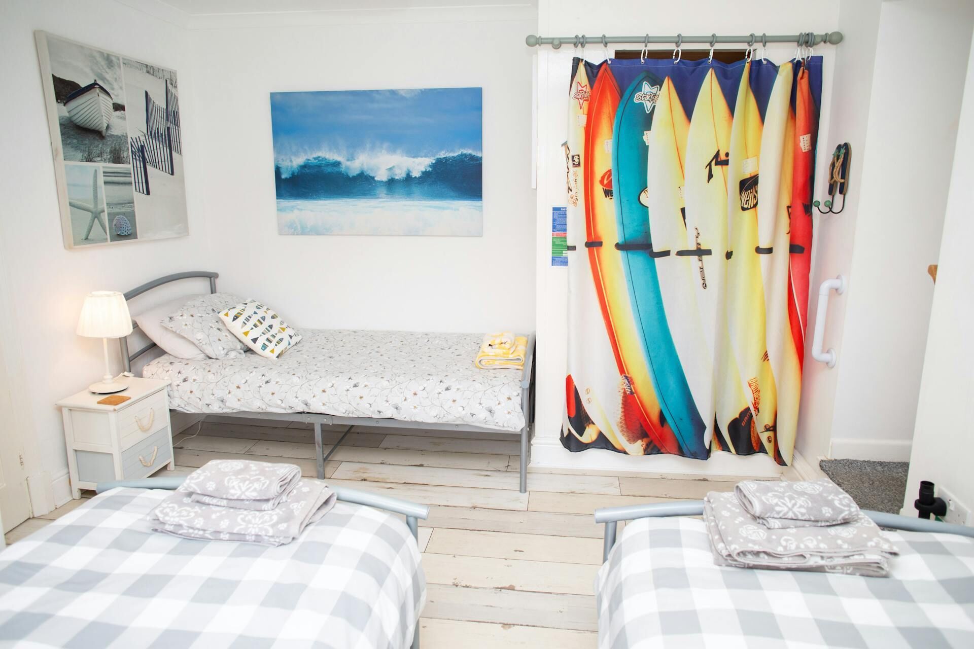 Ein Schlafzimmer mit zwei Betten, einem Nachttisch, einer Lampe, einem Wandbild und einem farbenfrohen Vorhang mit Surfbrett-Motiv.