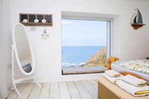 A bedroom with a large window overlooking the ocean, a bed with a patterned bedspread, a wooden bench with towels, and a white vanity with a mirror. - Brigydon, Tresaith - Two Bedroom Cottage, Sleeps 5 (Tresaith)