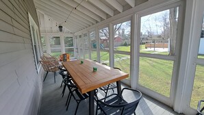 En overdækket terrasse med et træbord og stole, store vinduer og udsigt til baghaven.