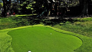 Un parcours de golf avec un green d’entraînement, des arbres et une aire de jeux en arrière-plan.