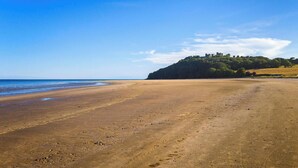 A wide sandy beach with a clear sky and a hill with structures in the distance. - 2 Rock Cottage, Llansteffan (Llansteffan)