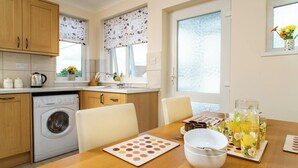 A kitchen with a washing machine, wooden cabinets, a dining table with a placemat, and a window with a view. - 2 Rock Cottage, Llansteffan (Llansteffan)