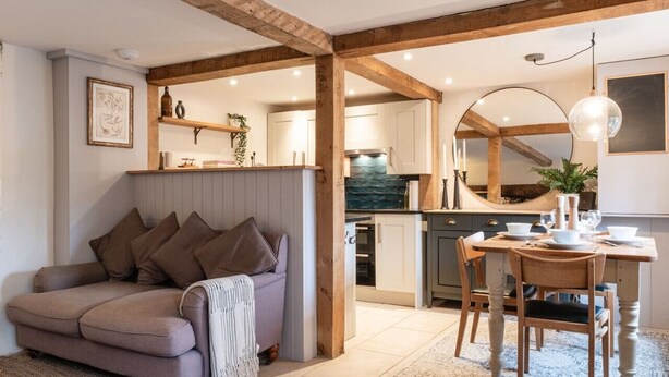 A cozy living room with a sofa, a dining table with chairs, and a kitchen area with wooden beams and white cabinets. - The Gamekeeper`s Cottage - Stunning 2 Bed (Leafield)