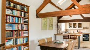 A dining area with a wooden table and chairs, a bookshelf filled with books, and a kitchen in the background.