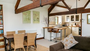 A spacious living room with a dining table, wicker chairs, a brown sofa, and a patterned throw pillow. A kitchen with wooden beams and white cabinets is visible in the background.