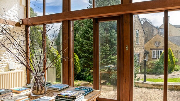 A room with a large window overlooking a garden, a wooden table with books, and a vase with branches. - Stunning Barn Conversion - Peaceful Retreat (Old Chalford)