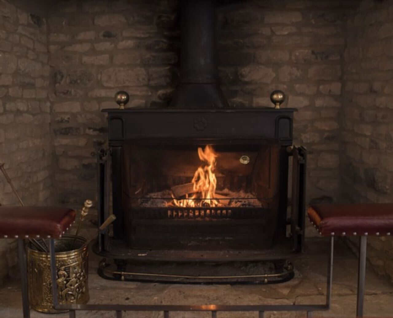 A wood-burning stove with a fire burning inside, flanked by two leather benches and a metal container on the floor.