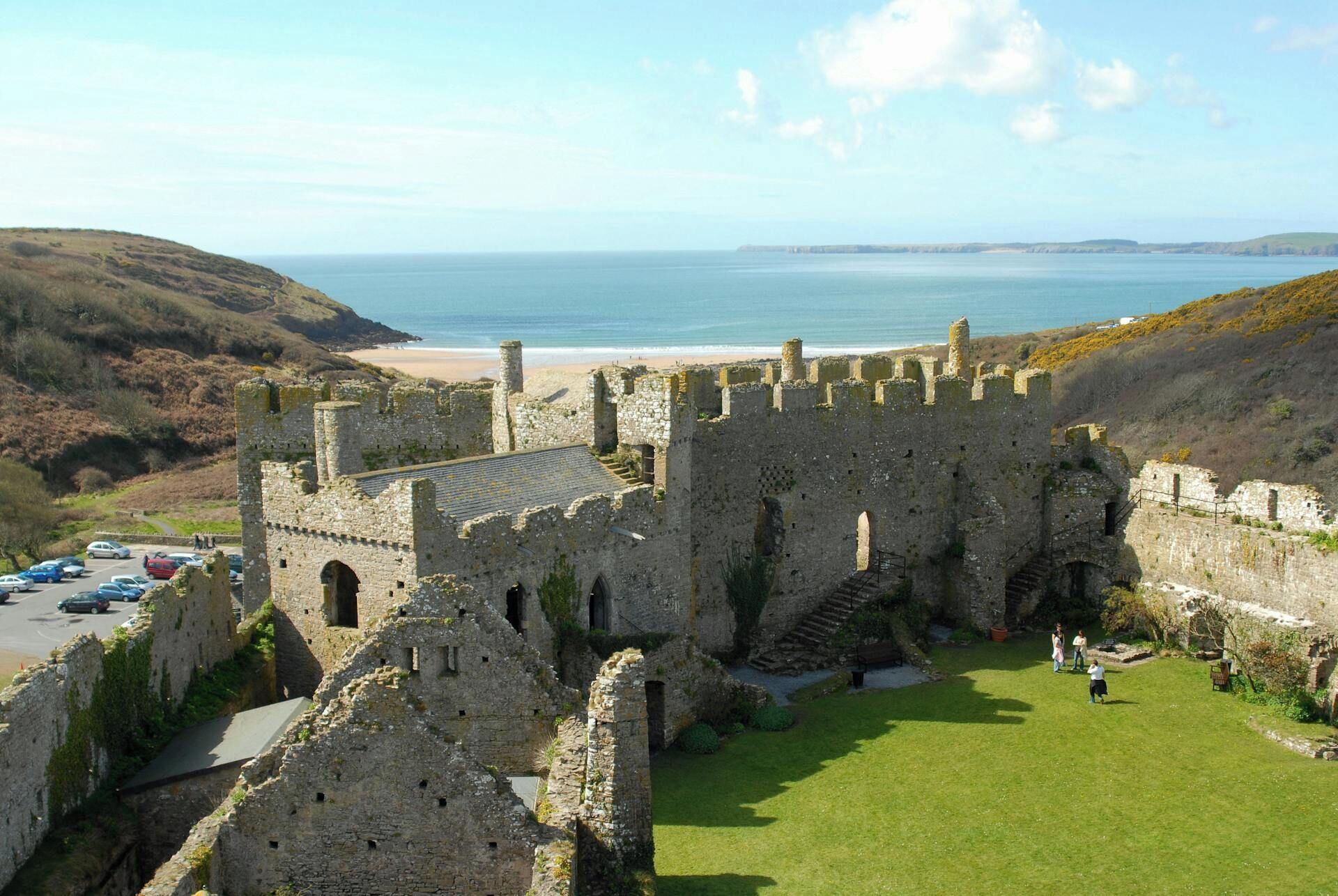 A historic castle with a grassy courtyard, stone walls, and a view of the beach and sea.