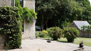 Un edificio in pietra con un cortile lastricato, circondato da vegetazione lussureggiante e una piccola costruzione con tetto spiovente.