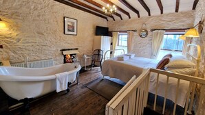 Une pièce rustique avec un mur en pierre, des poutres en bois, une baignoire blanche, un lit avec du linge de maison blanc et une fenêtre avec des rideaux.