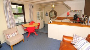 A cozy living room with a sofa, a dining table with red chairs, a kitchen area with a wooden countertop, and a large window with curtains. - 2 Castle Hall, Llangrannog (Llangrannog)