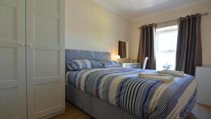 A bedroom with a bed, a bedside table, a lamp, a window with curtains, and a wardrobe. - Arwel, Aberporth - Three Bedroom Cottage, Sleeps 6 (Aberporth)