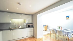 A modern kitchen with white cabinets, a microwave, and a sink. There is a dining area with a table and chairs, and a wicker chair. - Brodawel, New Quay - Three Bedroom Cottage, Sleeps 6 (New Quay)