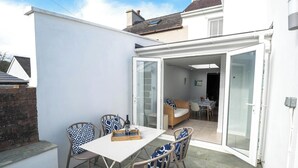 A patio with a table and chairs, a glass door leading to an interior room, and a brick wall. - Brodawel, New Quay - Three Bedroom Cottage, Sleeps 6 (New Quay)