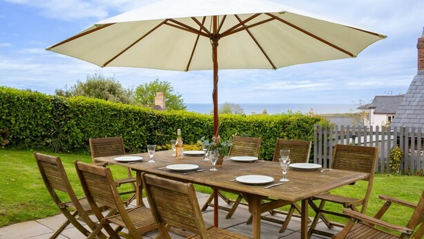 Une table en bois, dressée pour six personnes, protégée par un grand parasol et entourée de chaises en bois, dans un jardin agrémenté d’une haie et offrant une vue sur la mer.