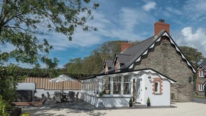 Un bâtiment en pierre avec une façade blanche, un grand espace extérieur aménagé avec des tables et des chaises, et une allée en gravier.