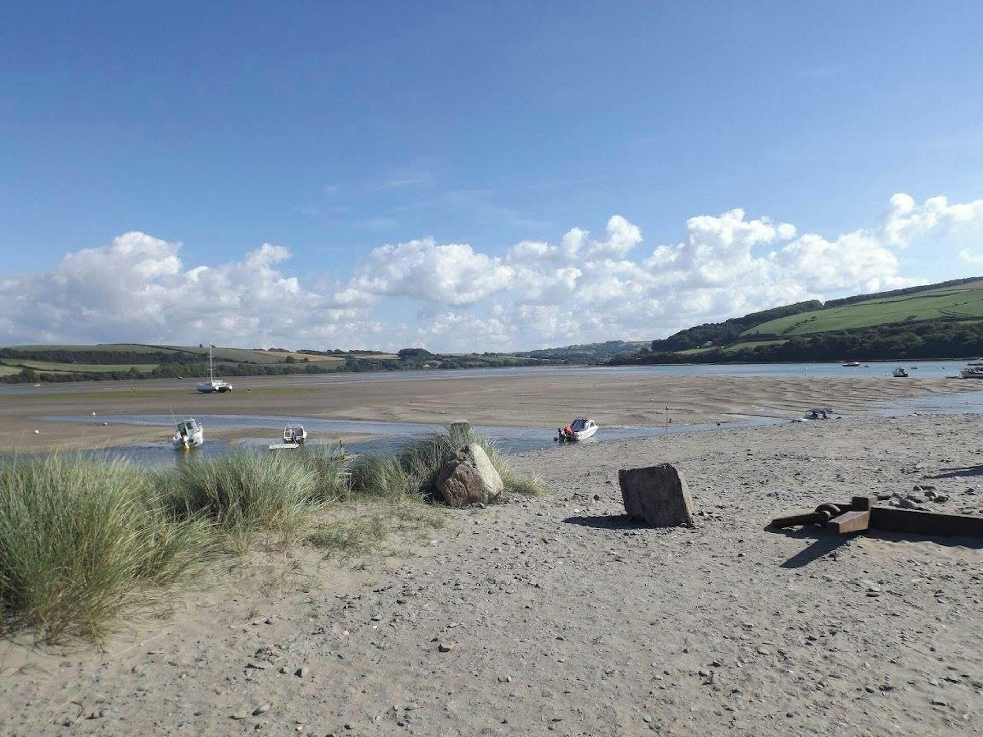 Un paysage côtier avec des bateaux sur une plage de sable, des zones herbeuses et des collines en arrière-plan.