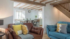Un salon avec un canapé en cuir marron et un canapé bleu, des poutres en bois au plafond, un coin repas à l’arrière-plan et une horloge au mur.