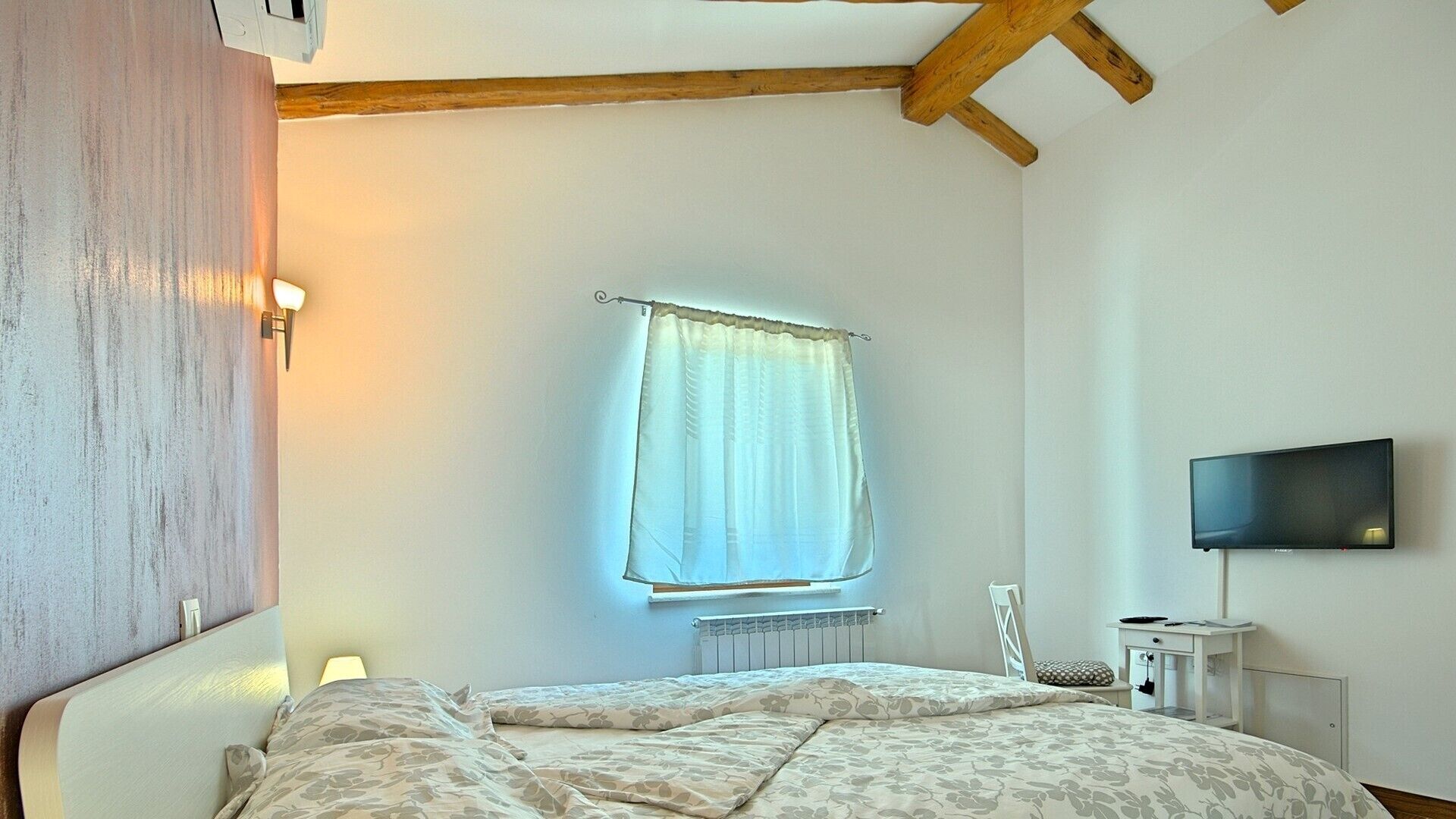 Een slaapkamer met een bed, een raam met gordijn, een radiator, een aan de muur bevestigde televisie en een stoel.