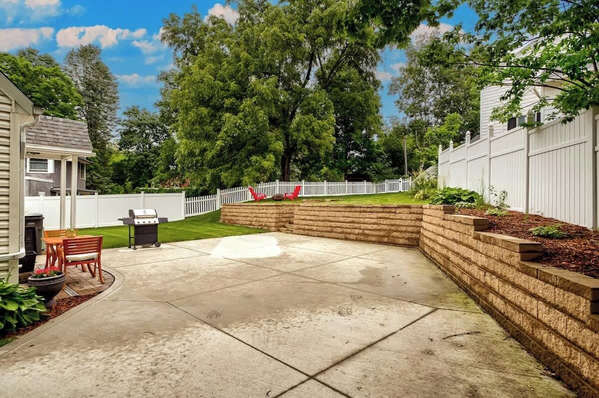 Un patio trasero con un porche, una parrilla, una valla blanca y un árbol.