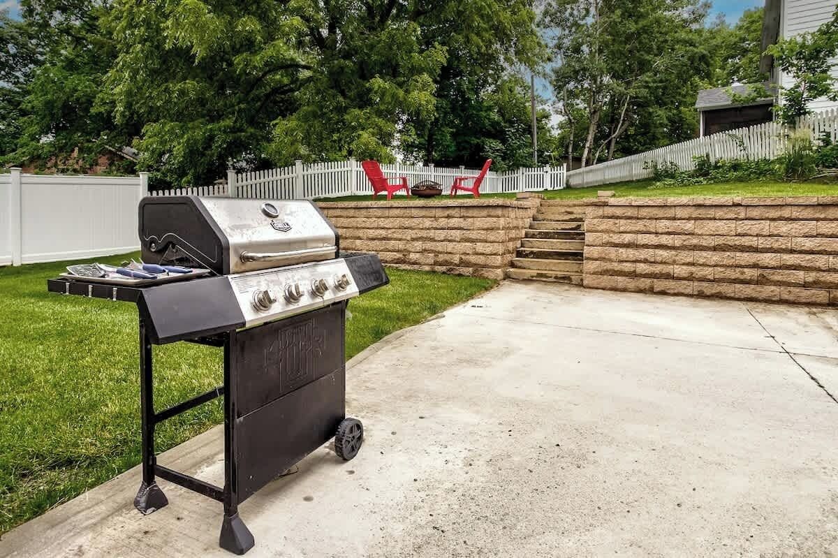 Een draagbare grill met een extra brander en een opbergvak, op een betonnen terras, met een witte hek en rode stoelen op de achtergrond.