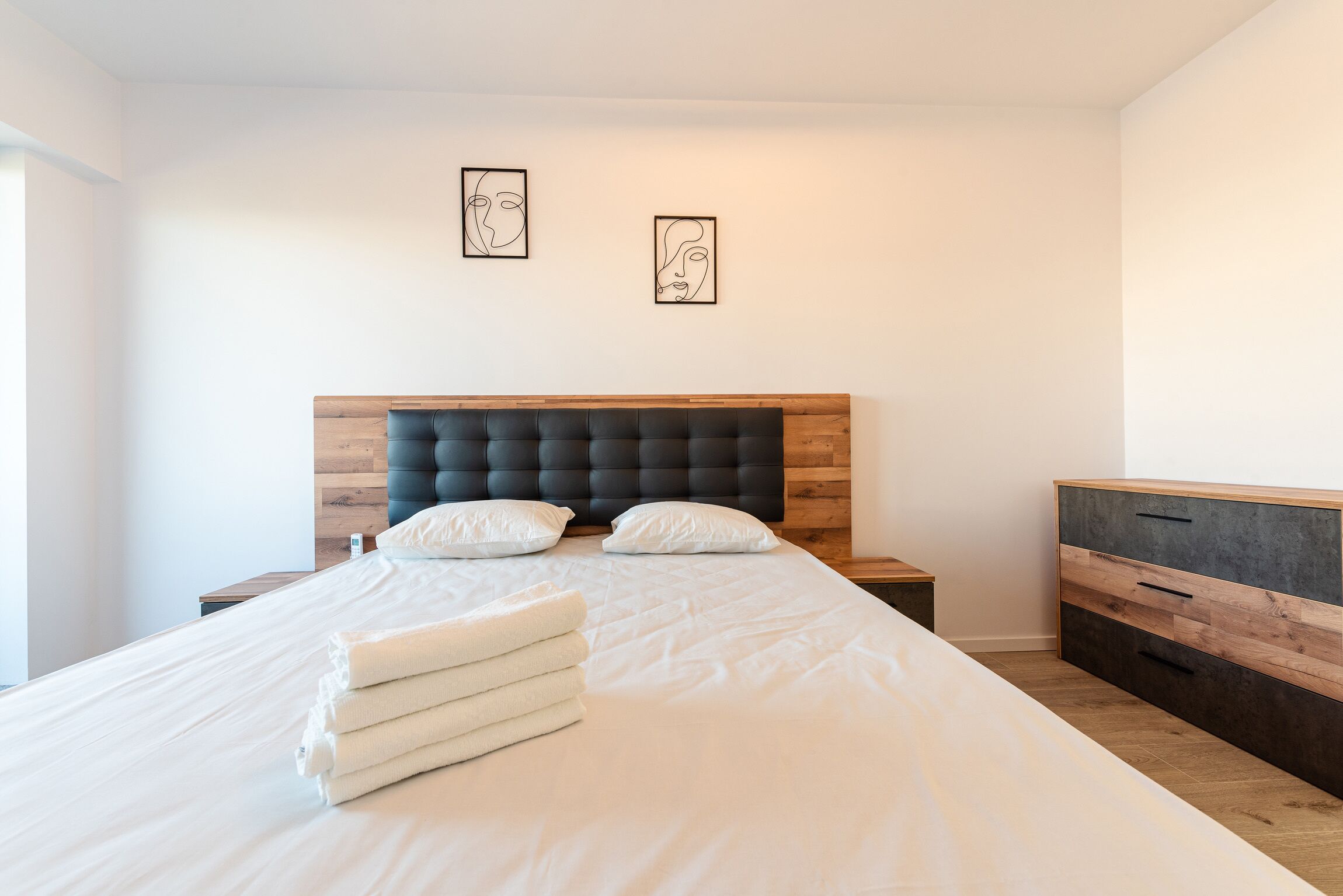 A neatly made bed with white linens and a stack of towels, a wooden headboard, and a nightstand with a lamp.