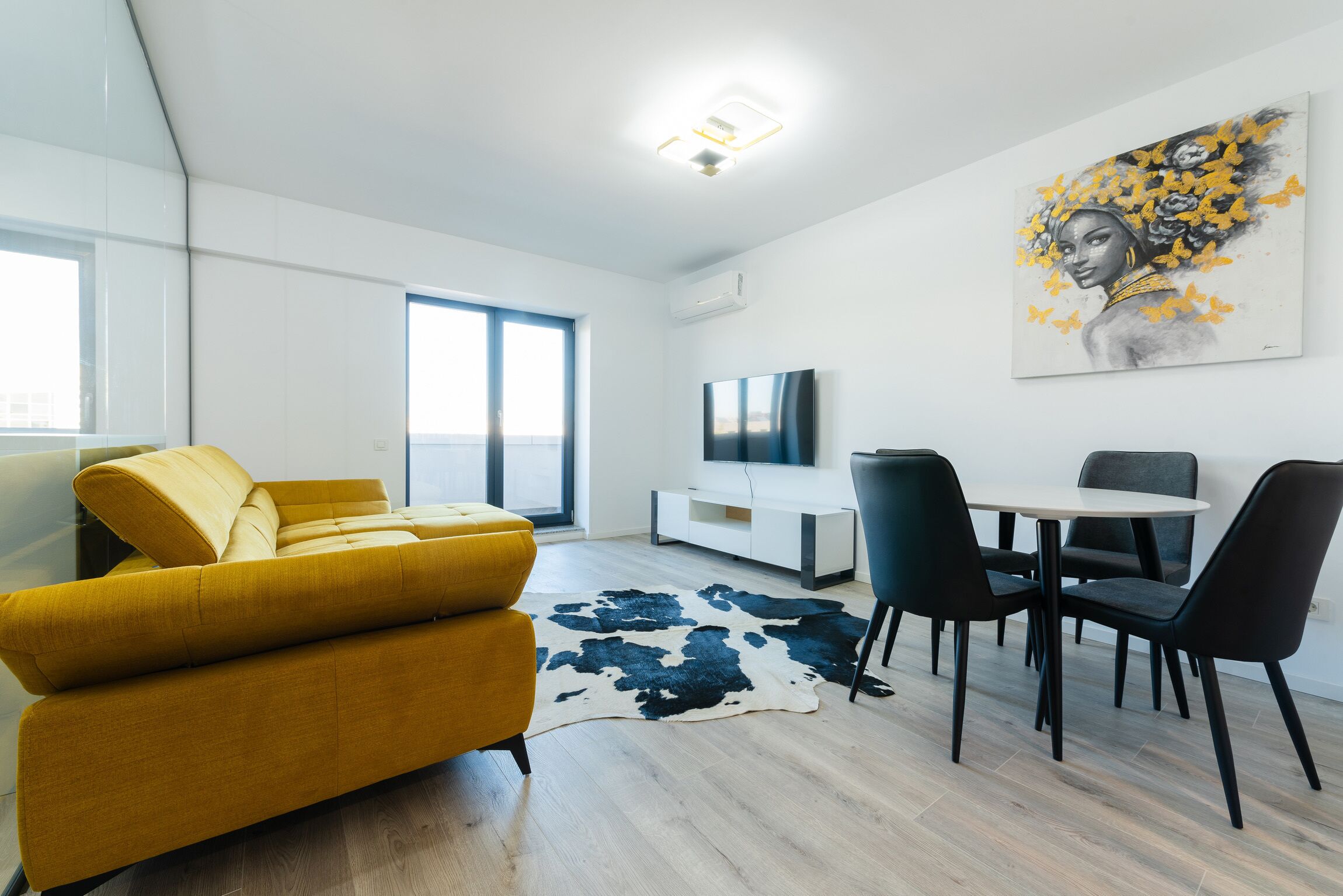Una sala de estar moderna con un sofá amarillo, una mesa de comedor con sillas, un televisor de pantalla plana y un cuadro grande en la pared.