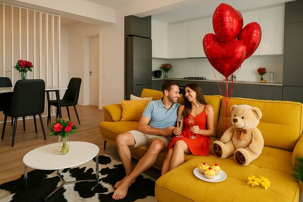 Un couple est assis sur un canapé jaune, un ours en peluche à leurs côtés, entouré de ballons rouges en forme de cœur. On aperçoit une table blanche avec un vase de fleurs, et au fond, une cuisine.