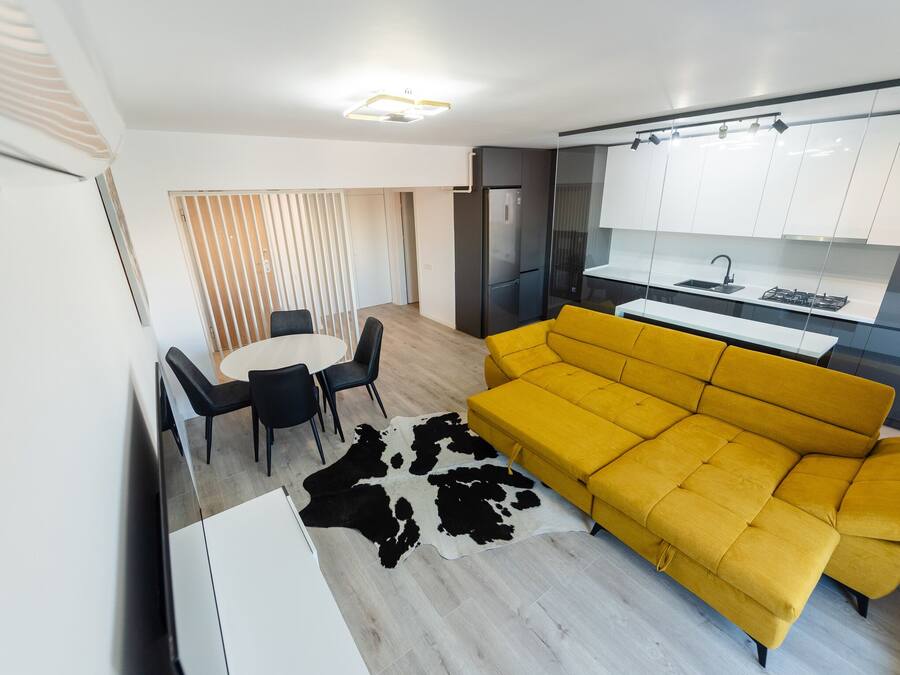 Una sala moderna con un sofá amarillo, un comedor con una mesa redonda y sillas, y una cocina con gabinetes blancos y un refrigerador negro.