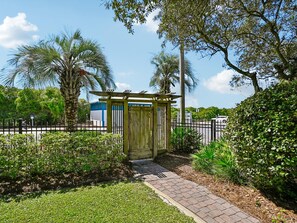 A gated entrance with a wooden pergola, surrounded by lush greenery and a paved pathway leading to a building with a blue and white facade.